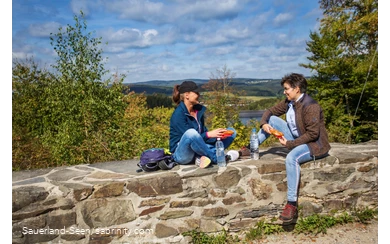 Zwei Frauen machen Rast auf einer Mauer mit Seeblick